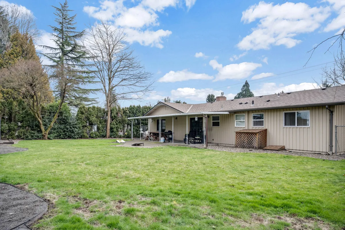 This image features a spacious backyard with a patio area, lush green lawn, and mature trees lining the property. The scene is set up for outdoor gatherings, gardening, and children playing, with plenty of open space for relaxation or energetic activities.