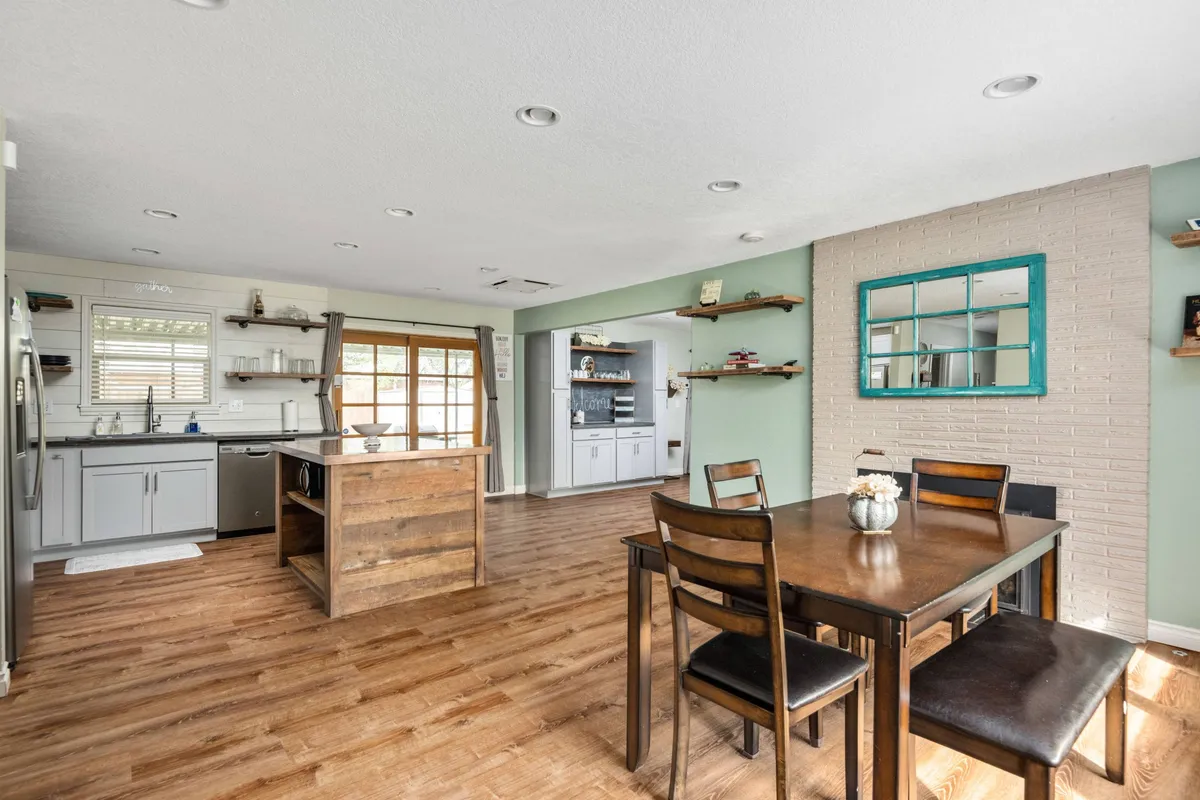 A welcoming view of the dining area with a table set for meals, adjacent to a kitchen island and illuminated by a large window. Decorative shelves add a curated touch, and the open setup reinforces the idea of a space designed for sharing meals and socializing.
