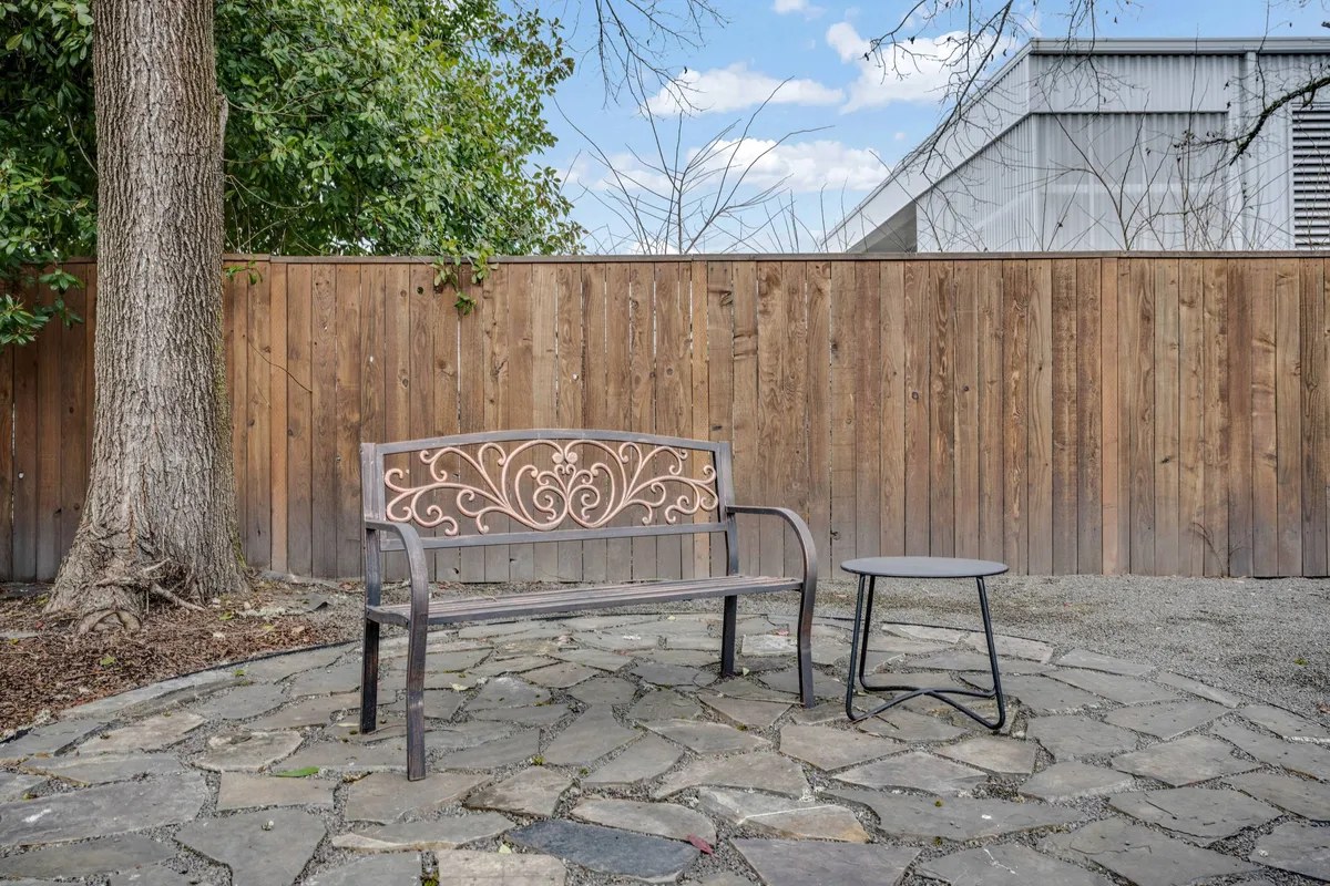 This photo captures a tranquil backyard scene with a decorative metal bench and a small round table set on a stone pathway. The space is framed by a wooden fence and shaded by a leafy tree, suggesting a private, comfortable area for relaxing outdoors.