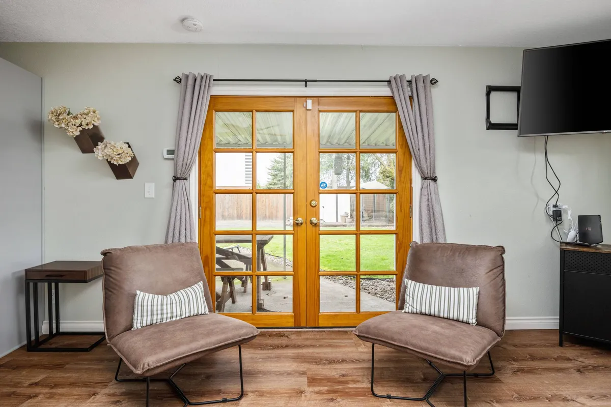A cozy lounge area with comfortable chairs, double glass doors that open to the backyard, and a TV mounted on the wall, with morning light streaming in and a clear view of the outdoor space.