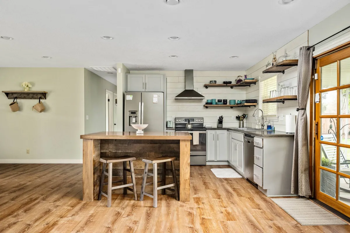 This image features a central wooden kitchen island surrounded by modern appliances and large windows that let in an abundance of natural light. The layout is open and airy, inviting both everyday family moments and larger gatherings. The scene captures the kitchen as a hub for conversation and culinary activity, with warm tones and contemporary style.
