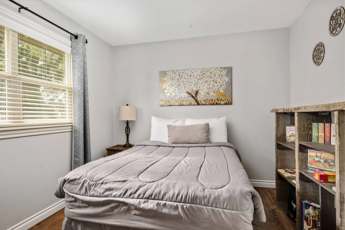 A serene bedroom featuring a bed with plush gray bedding, tree-themed artwork above the headboard, a nightstand with a softly glowing lamp, and a bookshelf with books and curated décor. The room exudes peacefulness and comfort, enhanced by inviting touches.