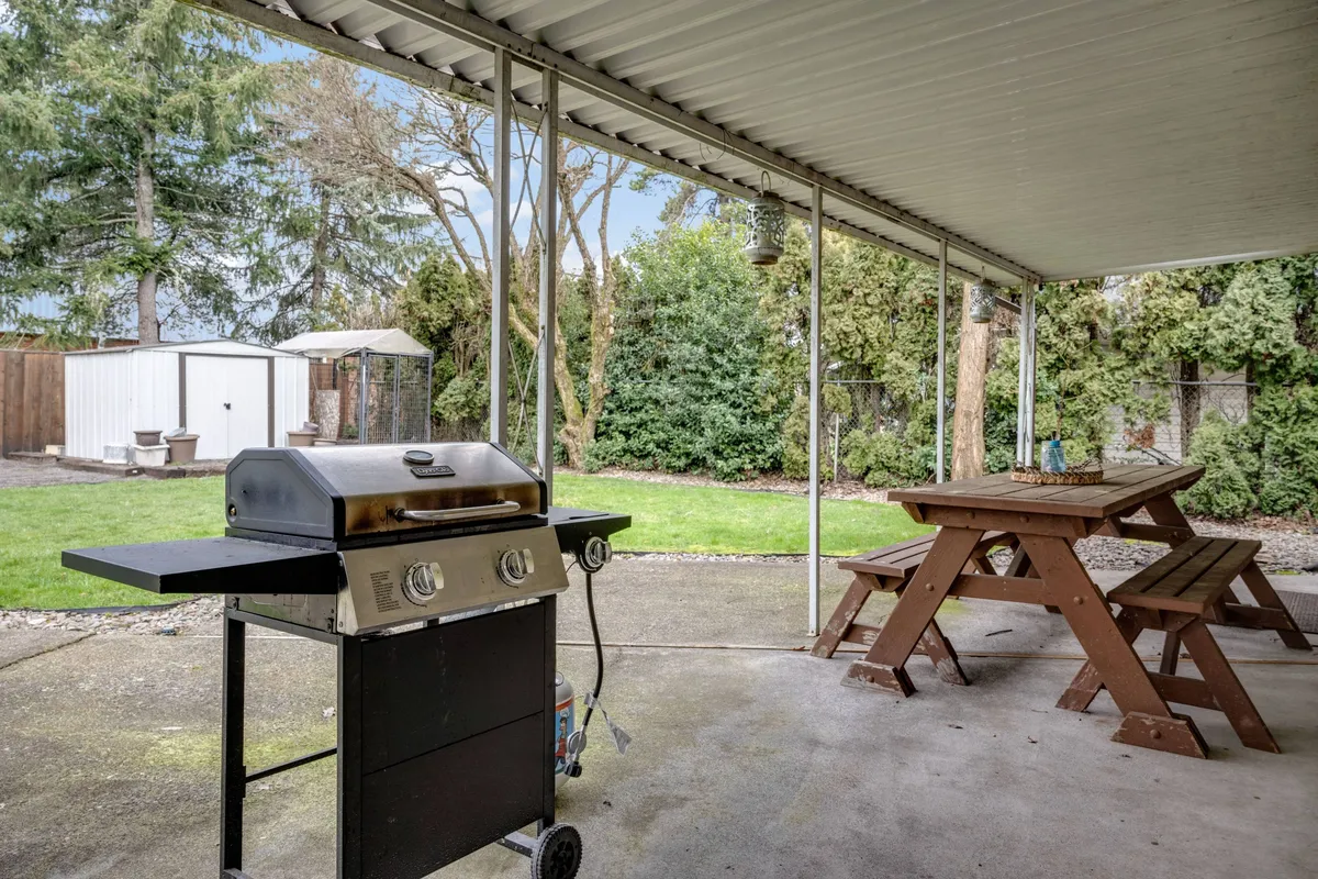 A backyard view featuring a grill, picnic table, and green lawn surrounded by hedges and a garden shed. The setup invites barbecuing, outdoor dining, and relaxed gatherings.