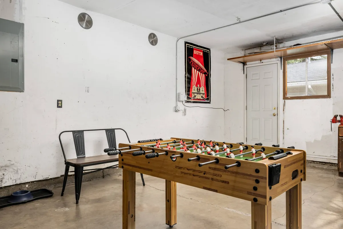 A dedicated recreation garage featuring a foosball table as the central element. The space includes playful decor, a poster on the wall, and a door that leads directly outside, suggesting easy access to the backyard. The overall vibe is casual and inviting, ideal for group games and leisure time.
