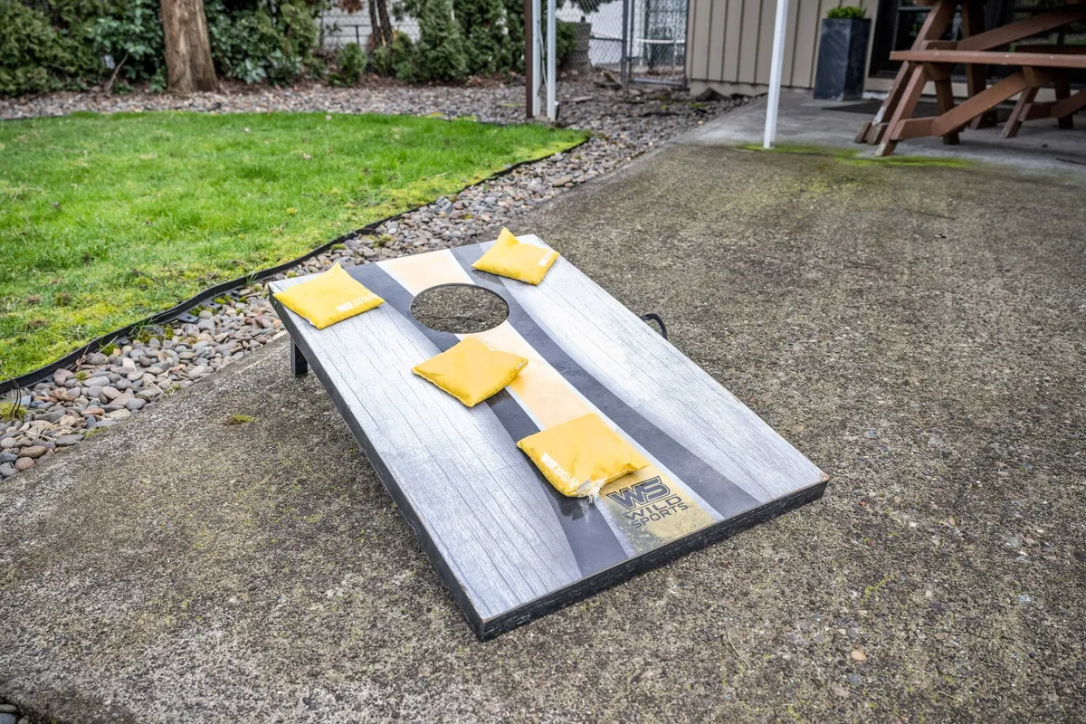 A vibrant backyard scene centered around a cornhole game set up on a lush green lawn. The scene conveys playfulness, community, and family fun in a private outdoor setting.