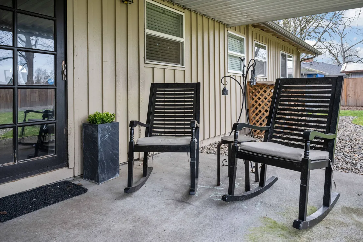 A serene exterior setting with rocking chairs and a potted plant on the porch, framed by a door and window. The calm and comfortable scene suggests a place to relax, socialize, or unwind after a busy day.