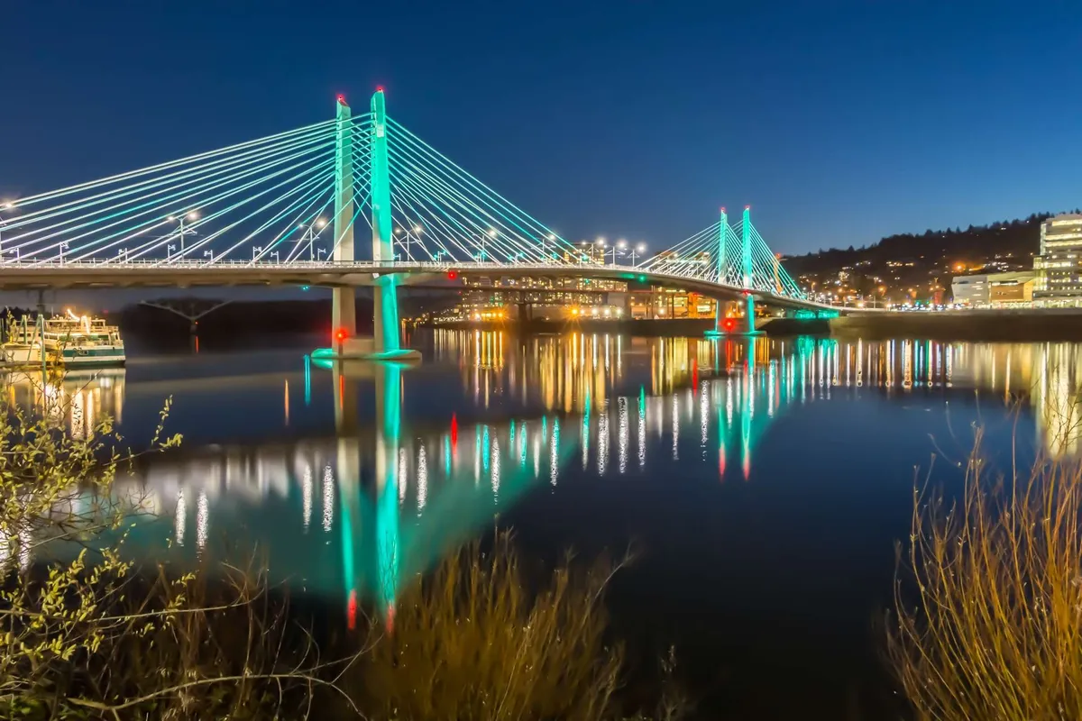 An inviting exterior scene featuring a picturesque bridge over water, with reflections and city lights creating an appealing backdrop. The location exudes a serene yet vibrant atmosphere, ideal for evening strolls and exploration.